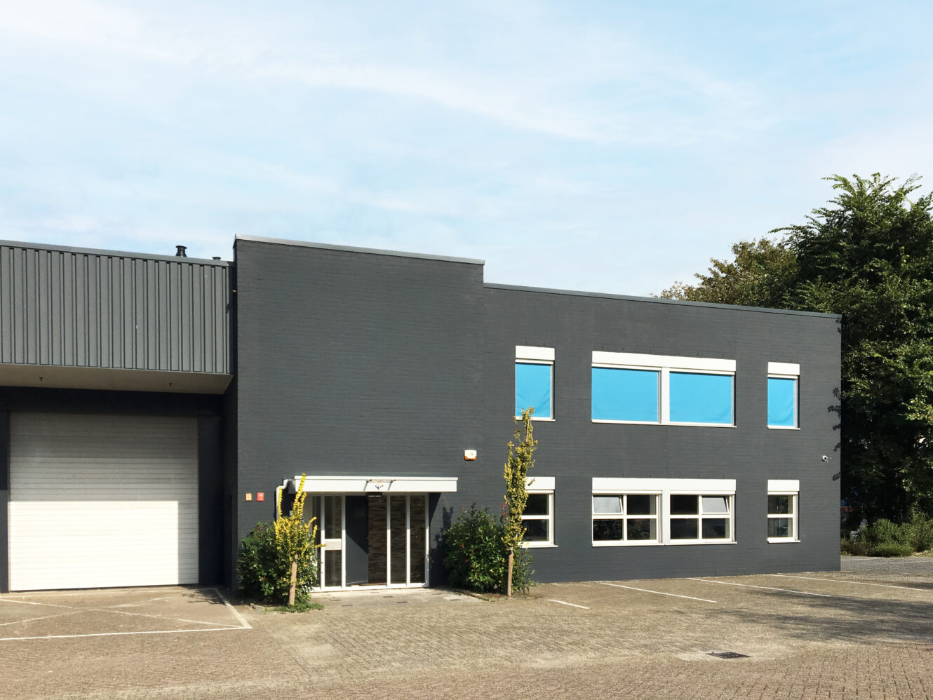 Een modern industrieel gebouw van twee verdiepingen met donkergrijze muren, grote ramen, een glazen toegangsdeur en een aangrenzende witte garagedeur. Het terrein aan de voorkant is bestraat en grotendeels leeg.