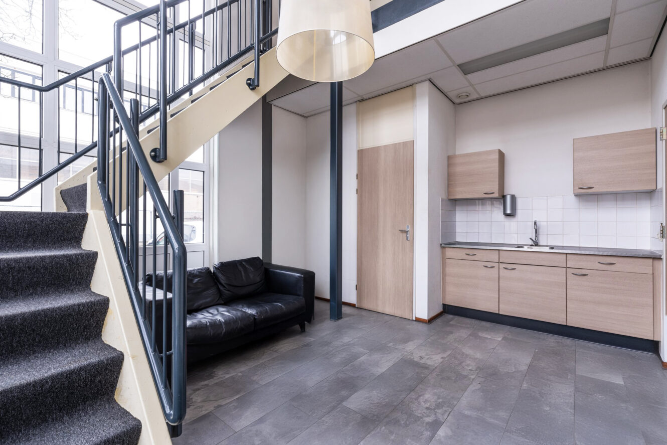 Modern interieur met een zwarte metalen trap, zwarte leren bank, kleine kitchenette met houten kastjes en tegelvloer. Grote ramen zorgen voor natuurlijk licht.
