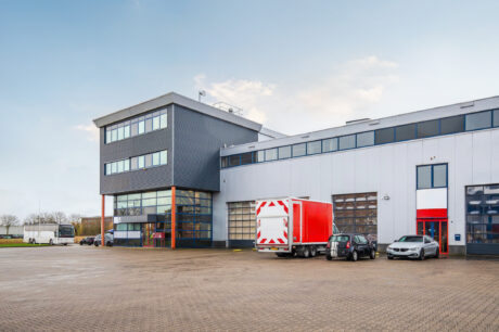 Modern industrieel gebouw met grote ramen, een rode bestelwagen en verschillende geparkeerde auto's op een ruim verhard terrein onder een heldere hemel.