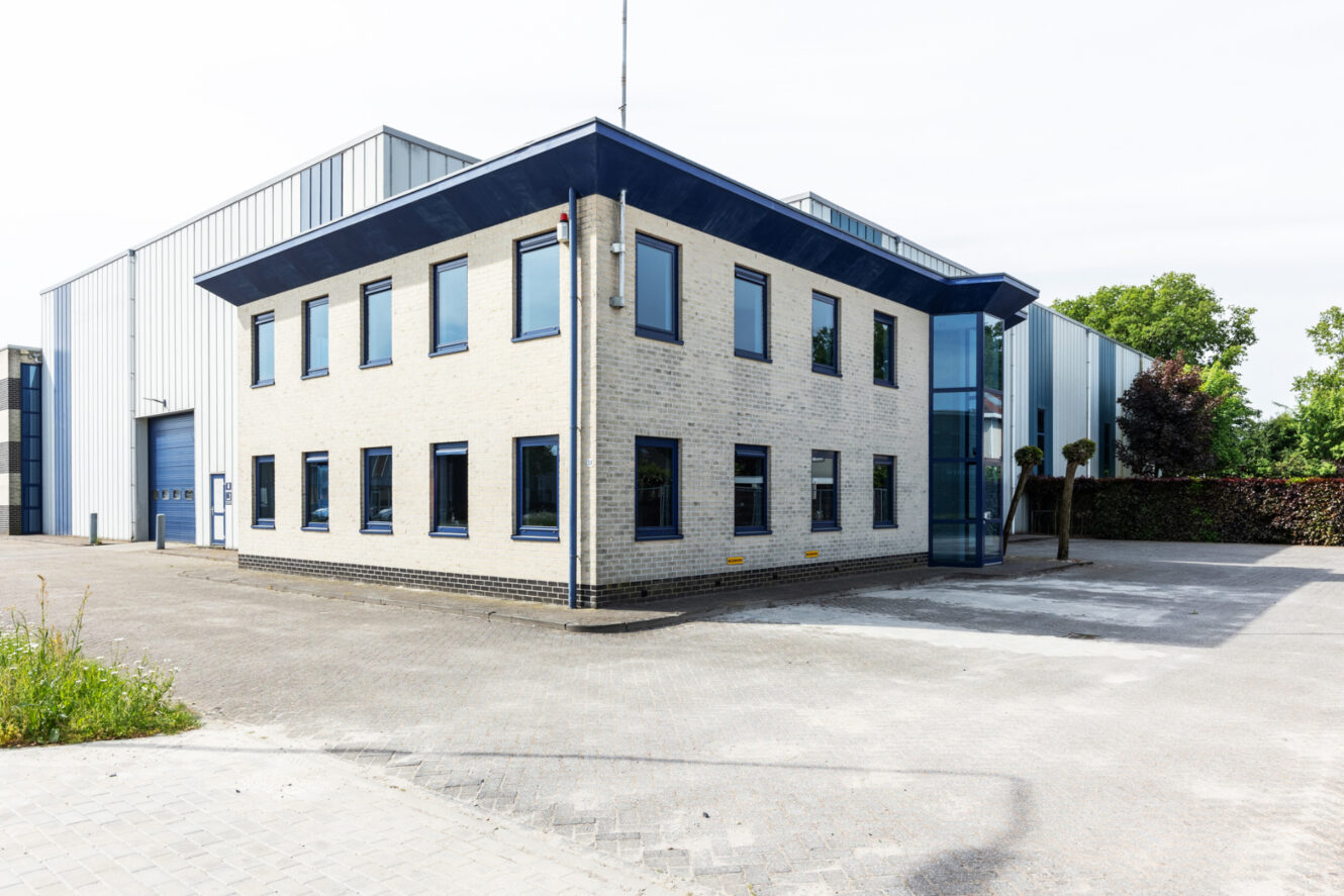 Een modern kantoorgebouw van twee verdiepingen met lichte bakstenen muren, blauwe kozijnen en een glazen ingang, omgeven door een geplaveide parkeerplaats.