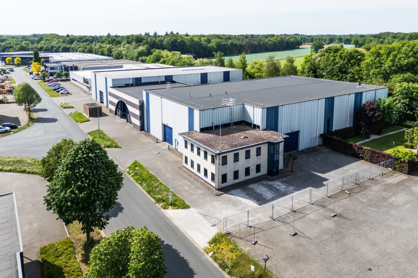 Luchtfoto van een industrieel complex met verschillende grote magazijngebouwen, een kantoorgebouw, parkeerplaatsen en omliggend groen.