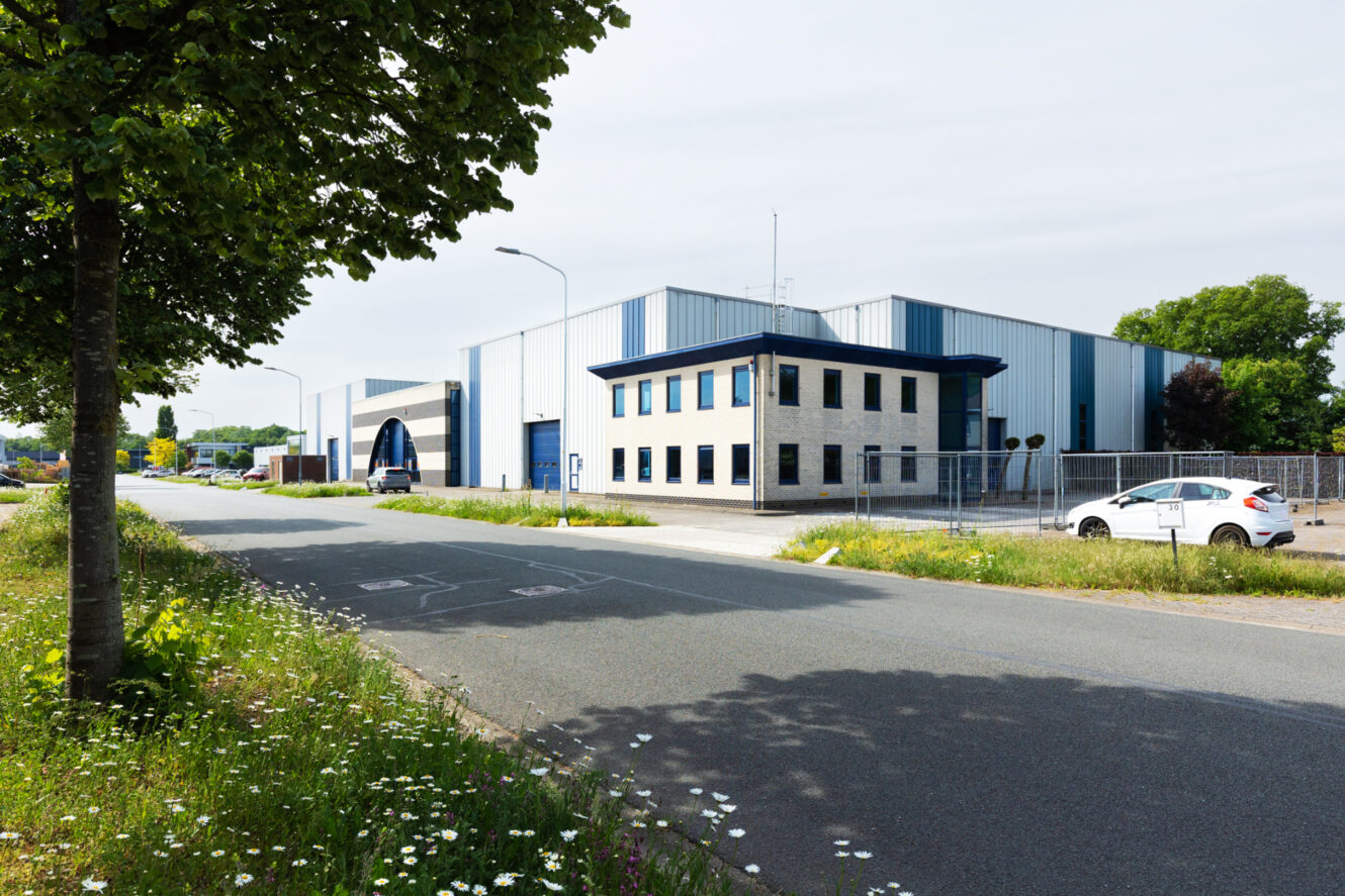 Een groot industrieel pakhuis met een klein kantoorgebouw ervoor, gelegen aan een rustige straat met geparkeerde auto's en bomen.