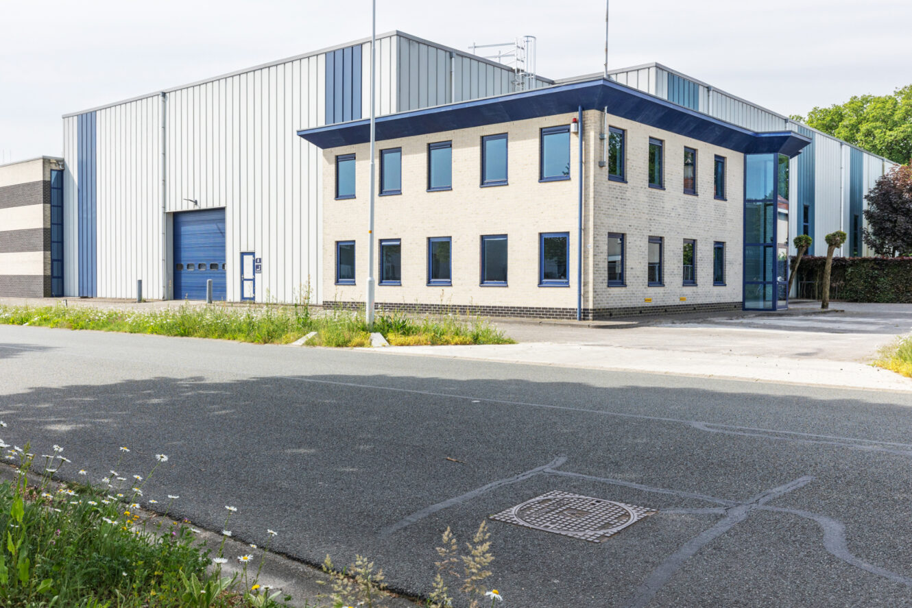 Modern industrieel gebouw met een blauw-witte buitenkant, grote ramen en een blauwe garagedeur, gelegen langs een verharde weg met wat groen in de buurt.