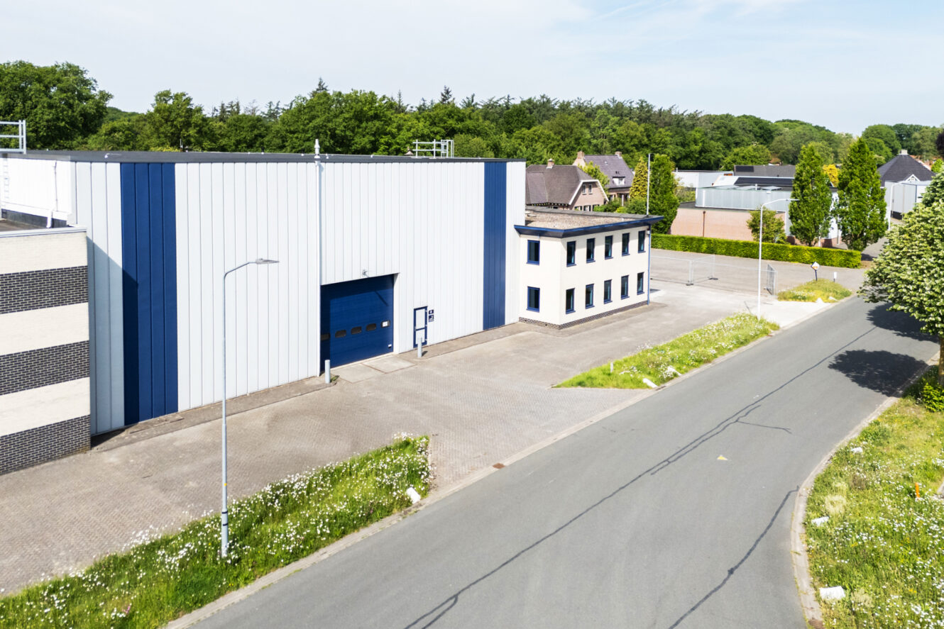 Een modern industrieel gebouw met een blauwe en witte gevel staat naast een rustige weg, omringd door groen en andere structuren.