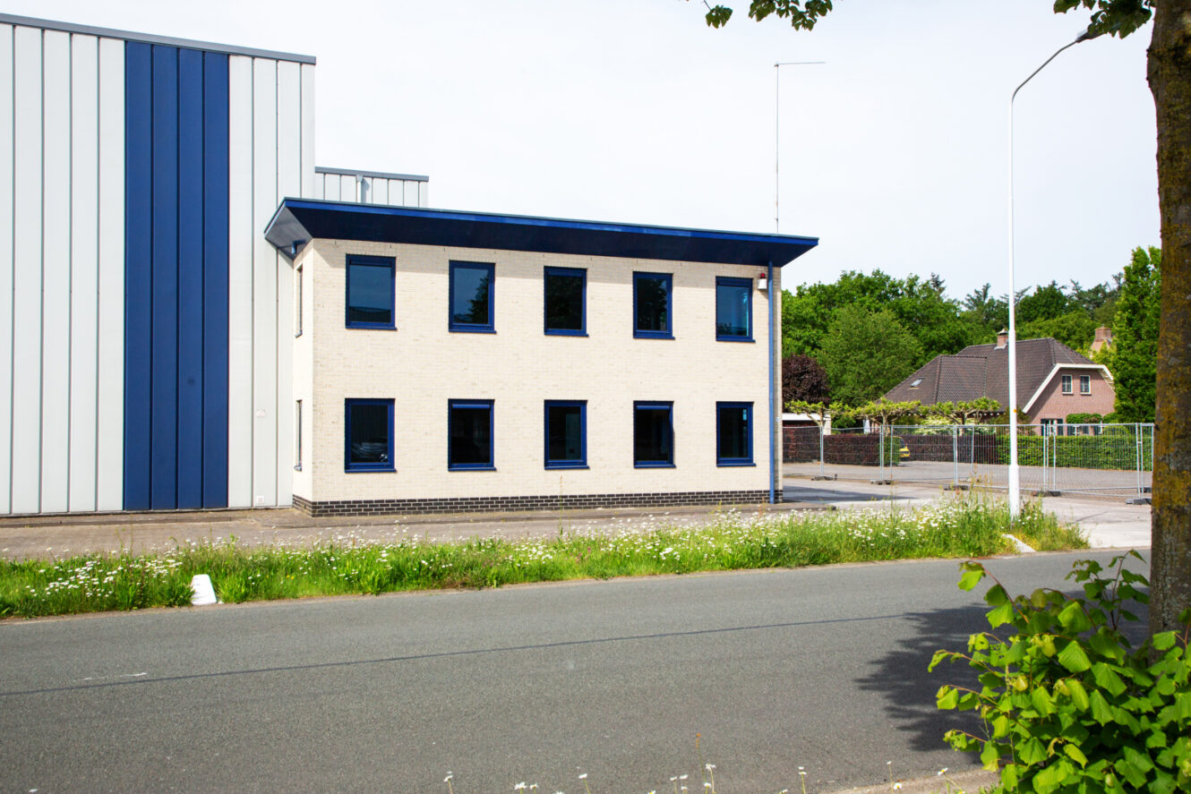 Een beige gebouw van twee verdiepingen met blauwe kozijnen staat langs een weg, naast een groter industrieel gebouw met verticale blauwe en grijze panelen. Bomen en huizen zijn zichtbaar op de achtergrond.