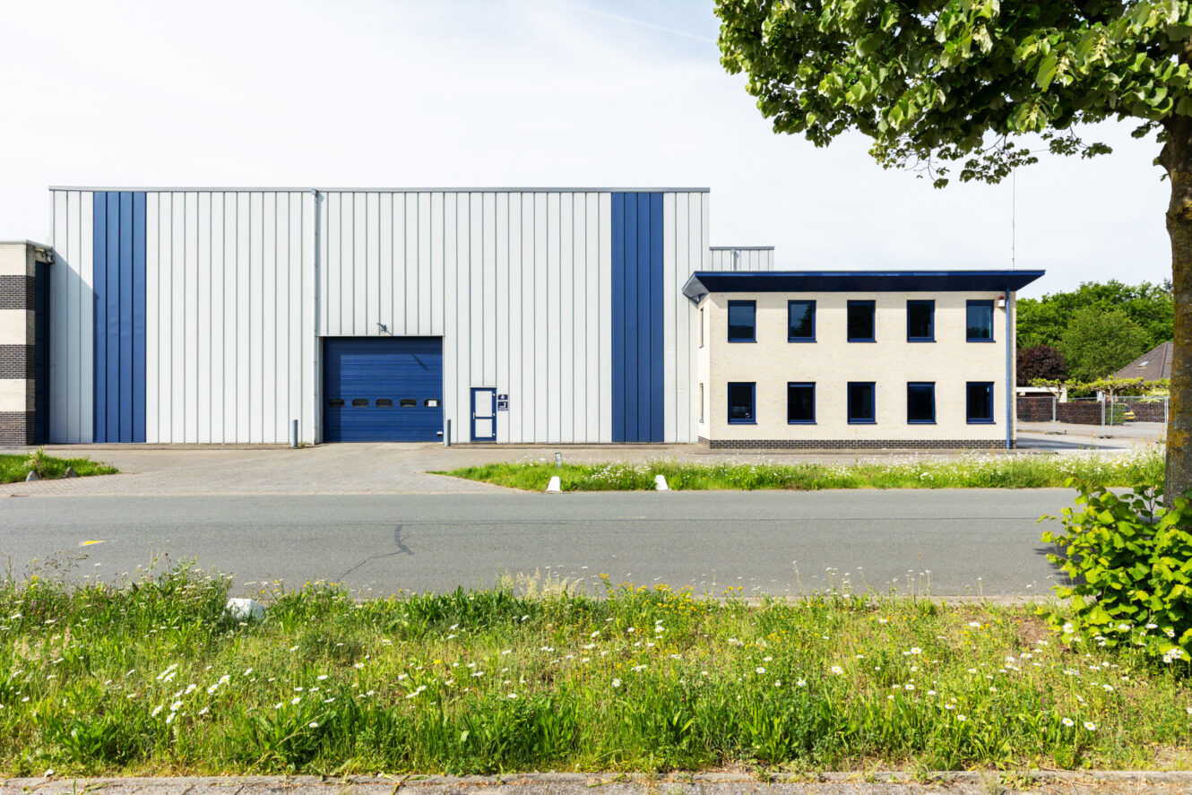 Een modern industrieel gebouw met een blauw-witte buitenkant, grote garagedeur, aangrenzend kantoorgedeelte en een boom op de voorgrond langs een weg.