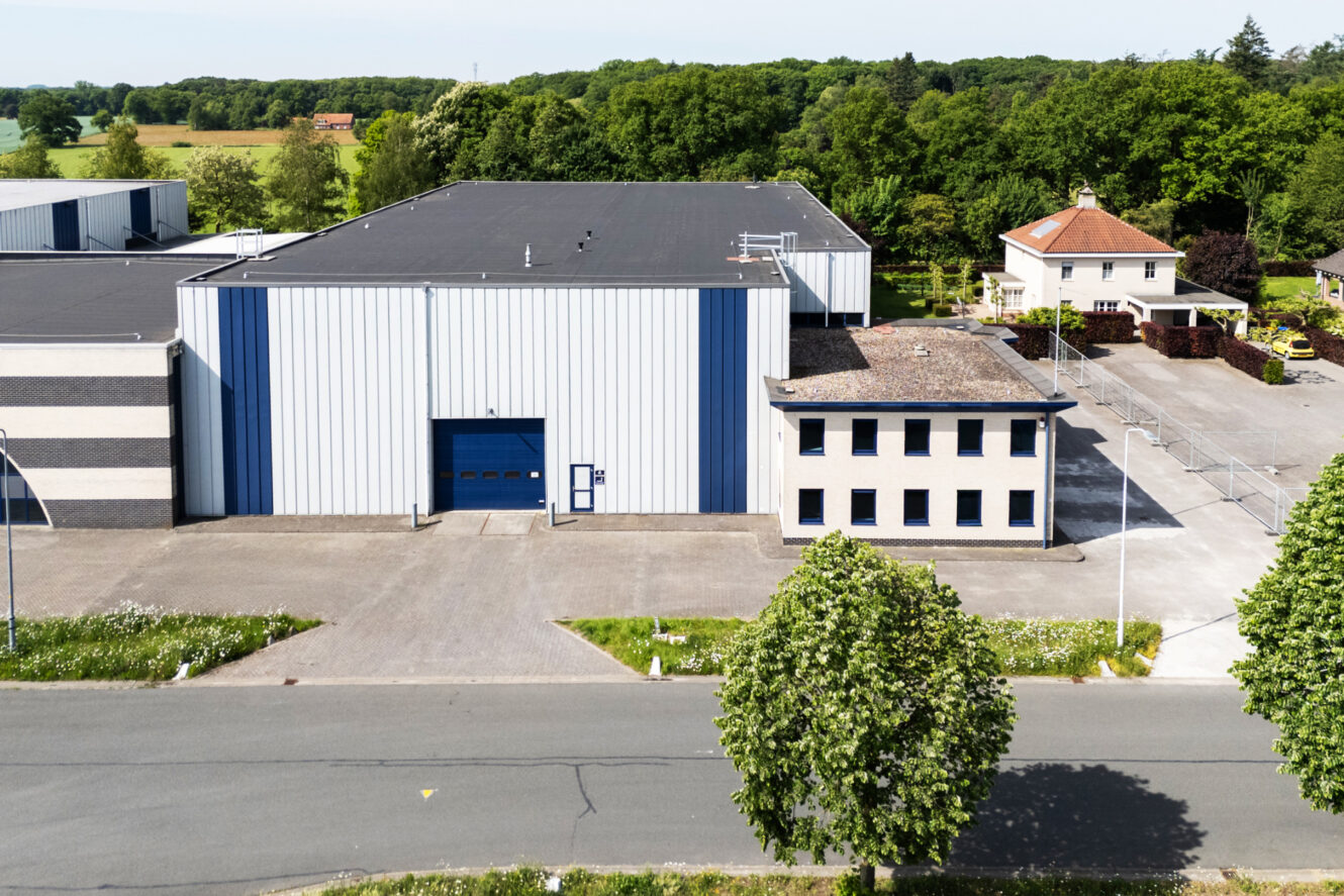Industrieel pakhuis met een blauwe en witte gevel, grote garagedeur en aangrenzend kantoorgebouw, gelegen bij een woonhuis en omringd door bomen.