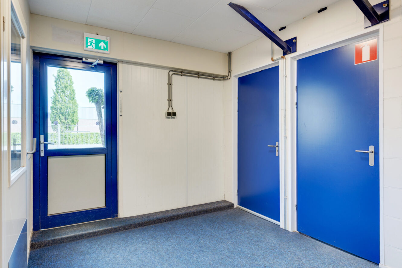 Een gang met blauwe deuren, een glazen deur met blauw frame, een bordje nooduitgang boven de deur en een rood bordje brandveiligheid aan de muur.