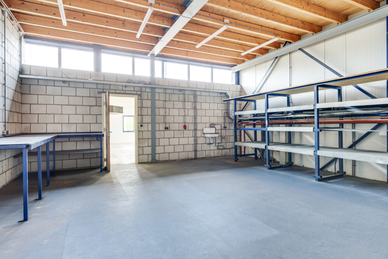 Leeg industrieel pakhuis met metalen rekken, werkbanken, zichtbare plafondbalken en een deur die naar een andere ruimte leidt. Natuurlijk licht valt binnen door de hoge ramen.