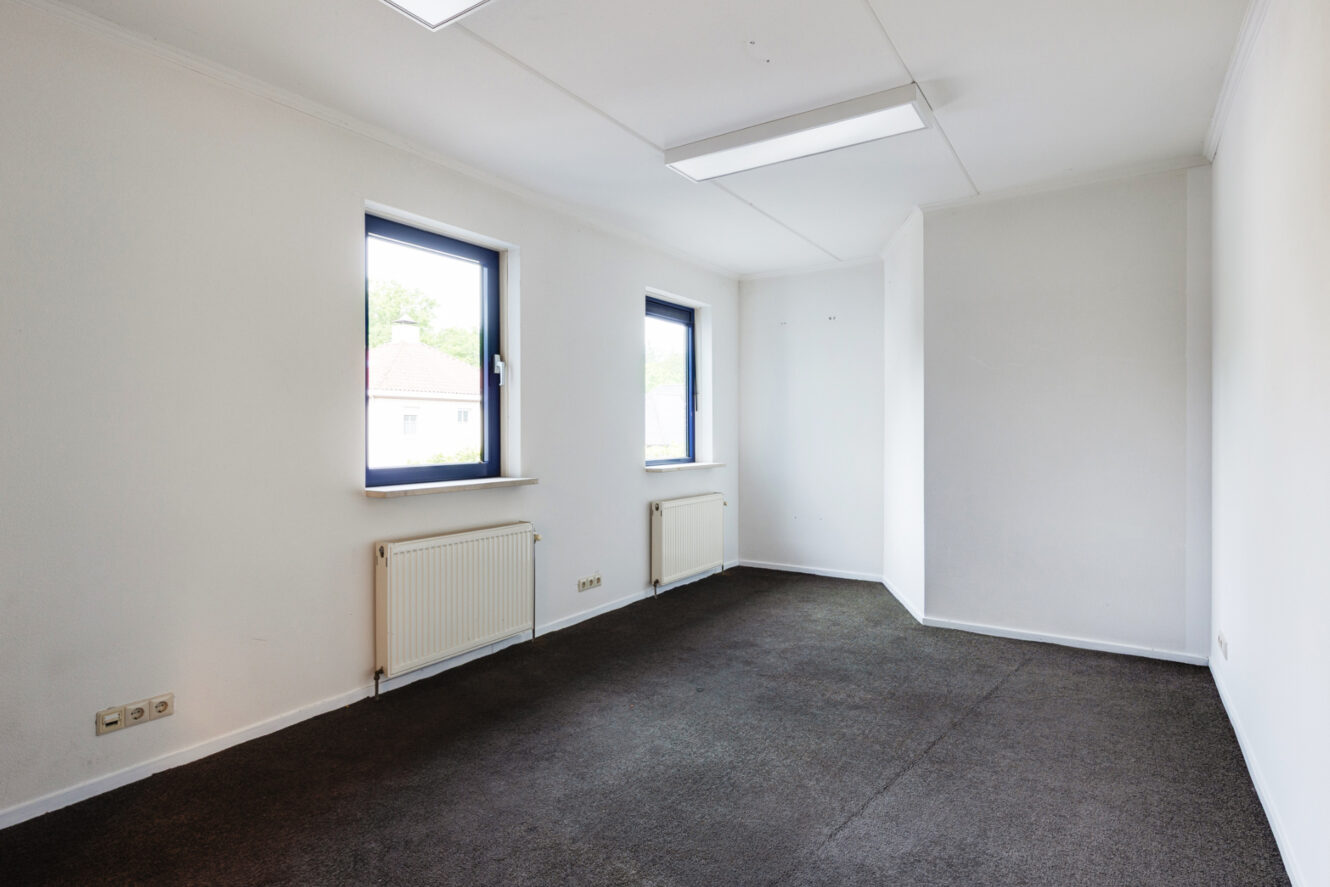 Een lege kamer met witte muren, twee ramen met radiatoren eronder en donkere vloerbedekking, verlicht door plafondverlichting.