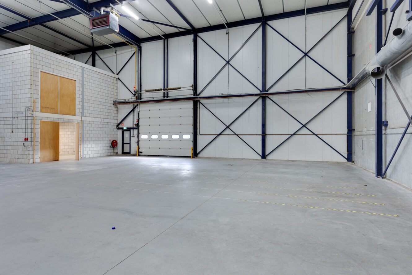 Interieur van een leegstaand industrieel pakhuis met hoge plafonds, betonnen vloeren, een grote garagedeur en een klein kantoor in een hoek.