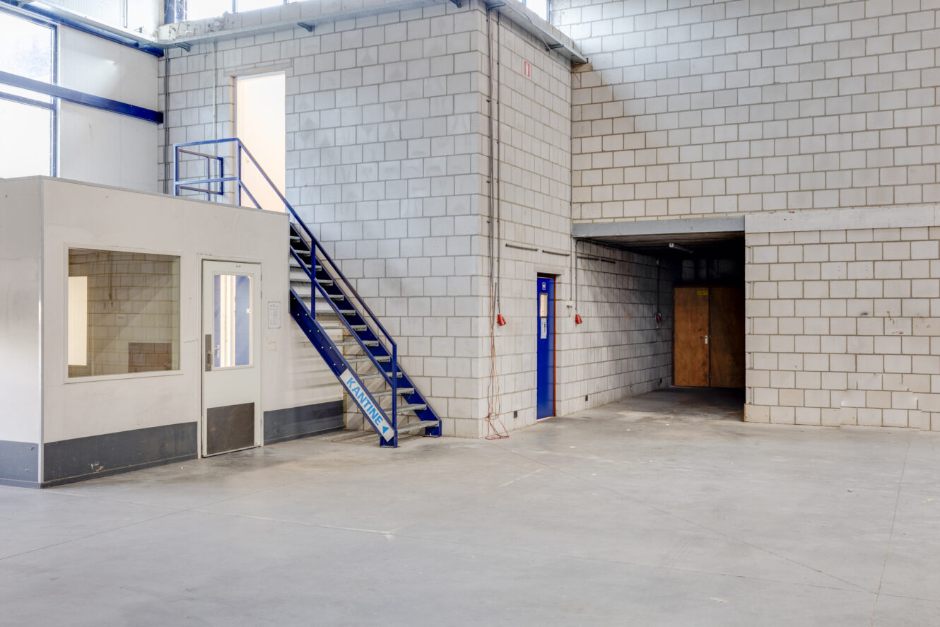 Interieur van een leeg industrieel pakhuis met muren van betonblokken, een klein kantoorgedeelte, een blauwe metalen trap en deuren die naar aparte kamers leiden.