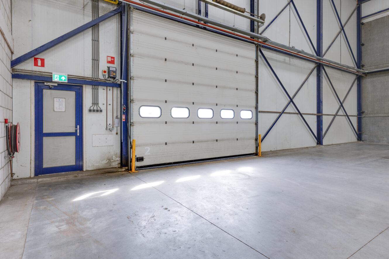 Interieur van een leeg pakhuis met een grote witte industriële garagedeur, een blauwe uitgangsdeur en betonnen vloeren; zonlicht schijnt door kleine ramen in de garagedeur.
