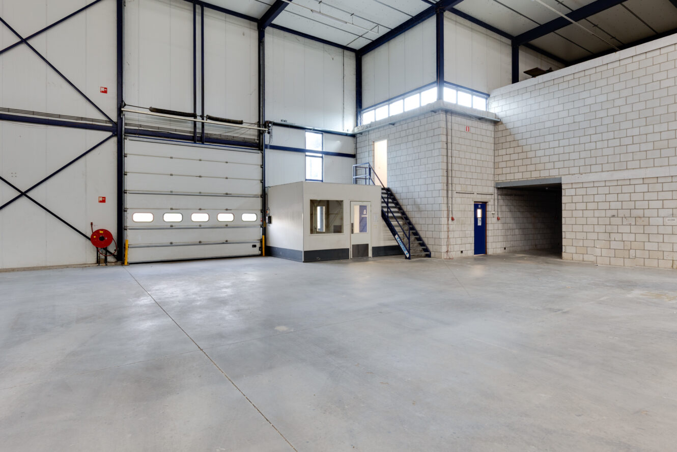 Groot, leeg pakhuis met betonnen vloeren, hoog plafond, witte bakstenen muren, een grote garagedeur, een klein kantoor en een trap naar een tweede verdieping.