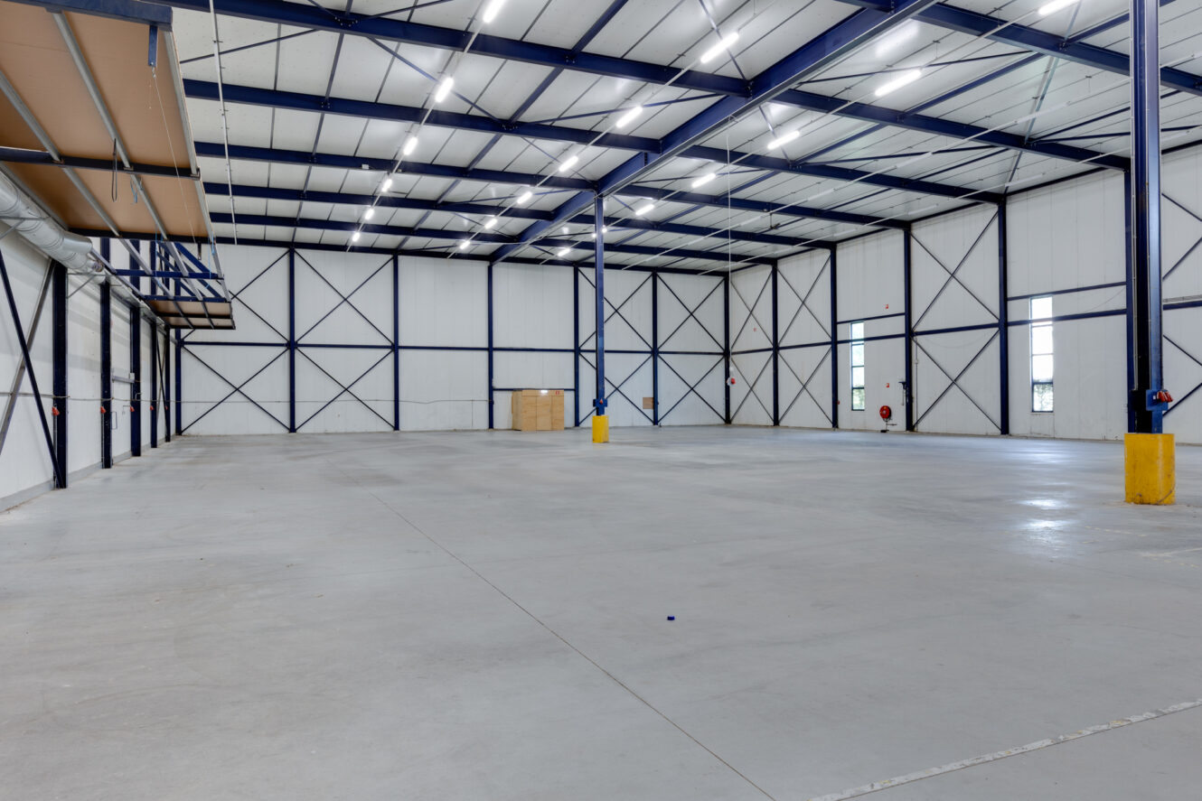 Ruim leeg magazijninterieur met hoge plafonds, heldere verlichting, betonnen vloer, metalen balken en een paar verspreide voorwerpen tegen de muren.