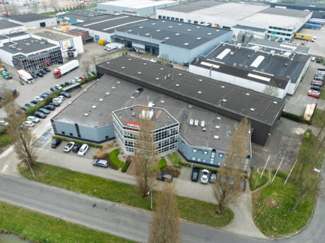 Luchtfoto van een industrieel gebouw met parkeerplaats, omringd door bomen en andere magazijngebouwen.