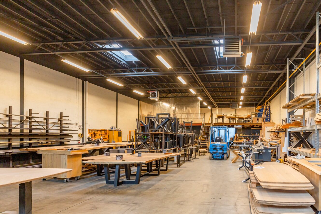 Ruime werkplaats met houten tafels, planken, gereedschap, metalen rekken en industriële verlichting, met een mezzanine en georganiseerde werkplekken.