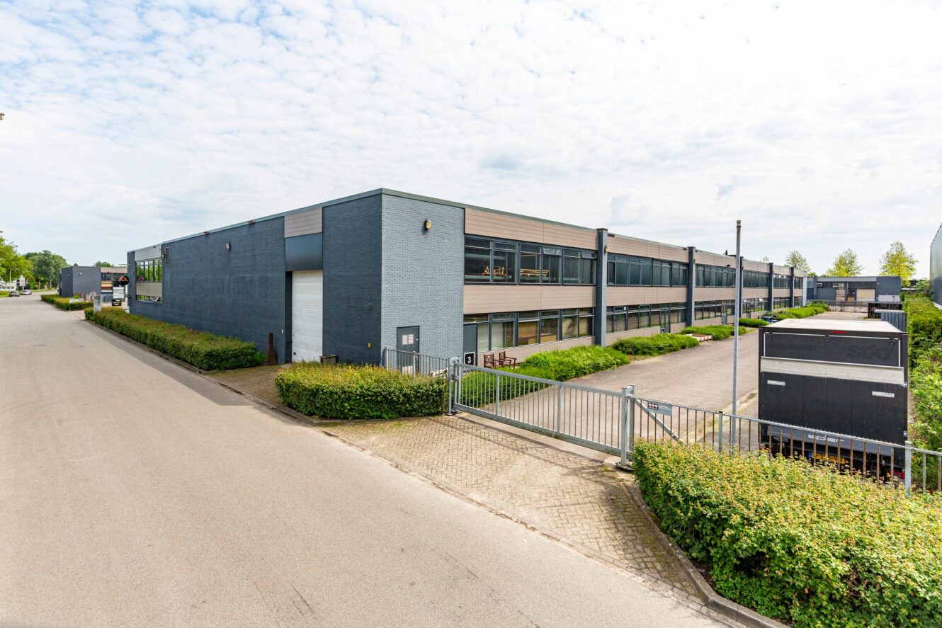 Een modern industrieel pakhuis met grijze muren, grote ramen en een omheinde ingang, gelegen aan een rustige straat met veel groen.