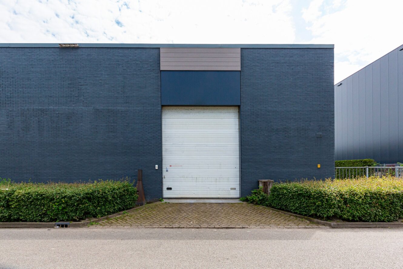 Een grijs industrieel gebouw met een grote gesloten witte garagedeur, omringd door lage groene struiken en een verharde oprit.