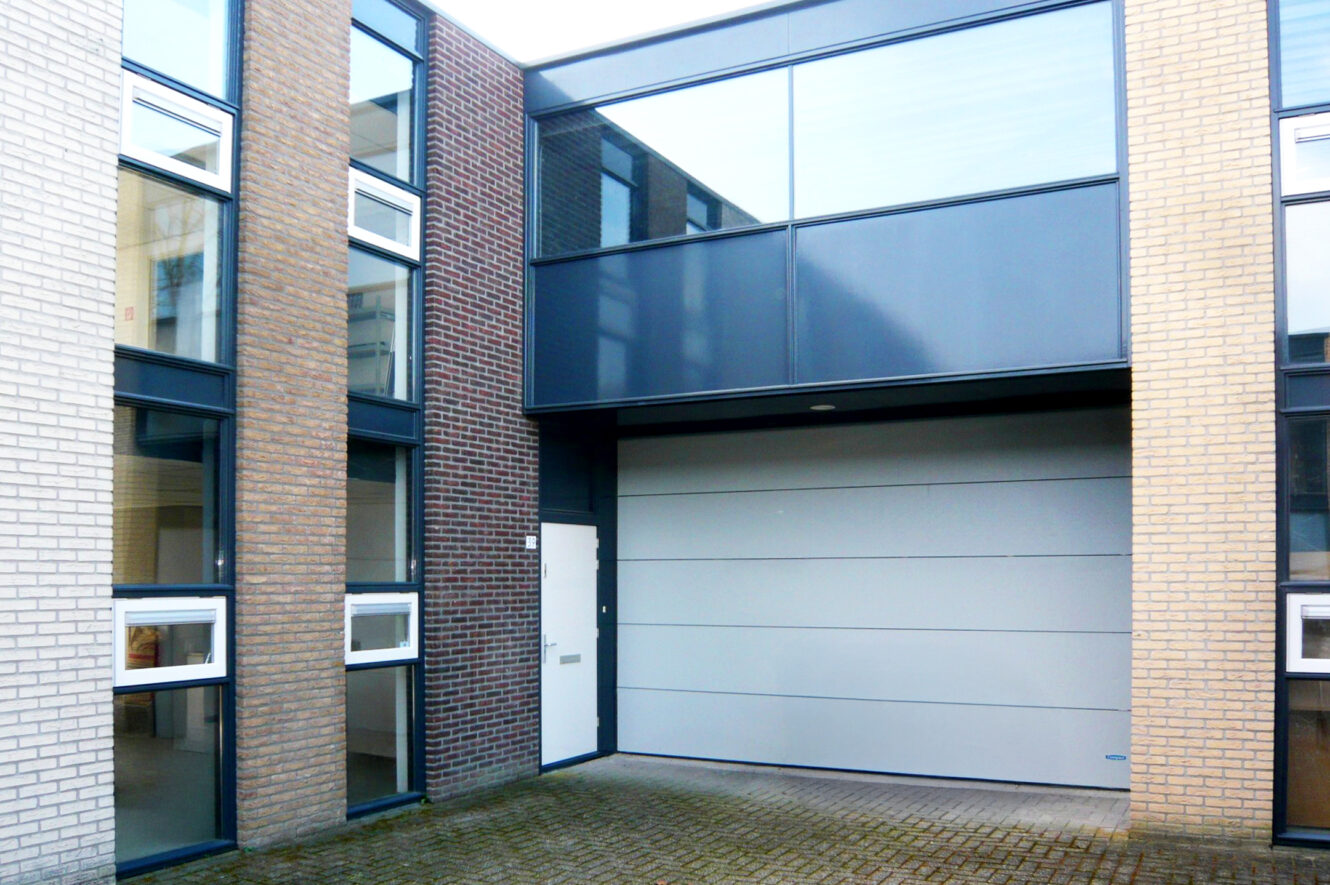 Moderne gebouwgevel met drie soorten metselwerk, grote glazen ramen, een witte deur en een brede grijze garagedeur op een geplaveide binnenplaats.