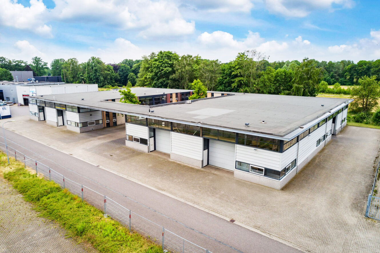 Een modern industrieel magazijncomplex met platte daken en grote garagedeuren, omgeven door een geplaveide binnenplaats en groen op de achtergrond.