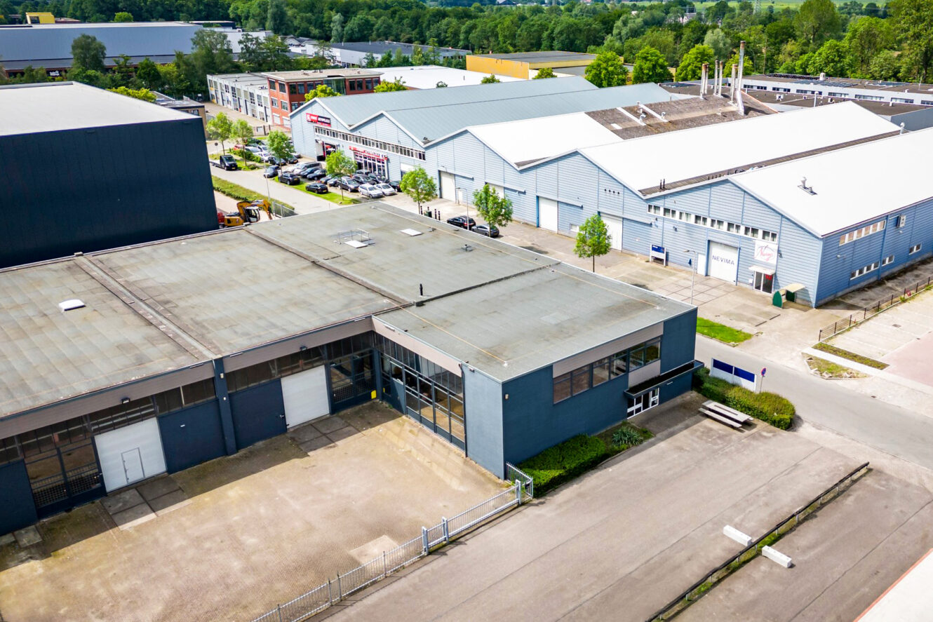 Luchtfoto van verschillende industriële pakhuizen met parkeerplaatsen en omliggend groen op een heldere dag.