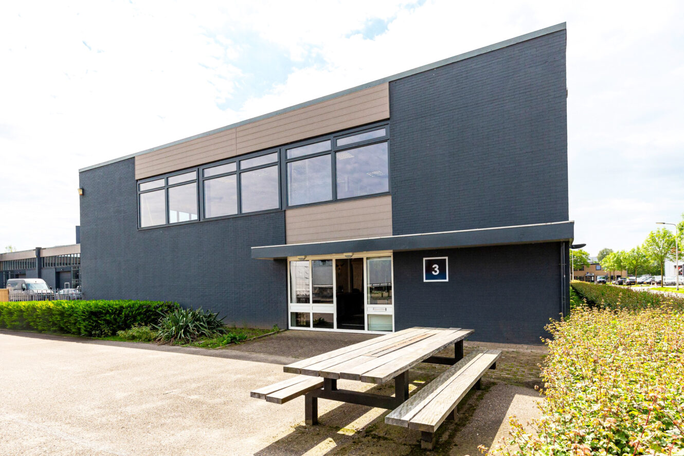 Een modern commercieel gebouw van twee verdiepingen met grote ramen, een bord met nummer 3 bij de ingang en houten picknicktafels aan de voorkant.