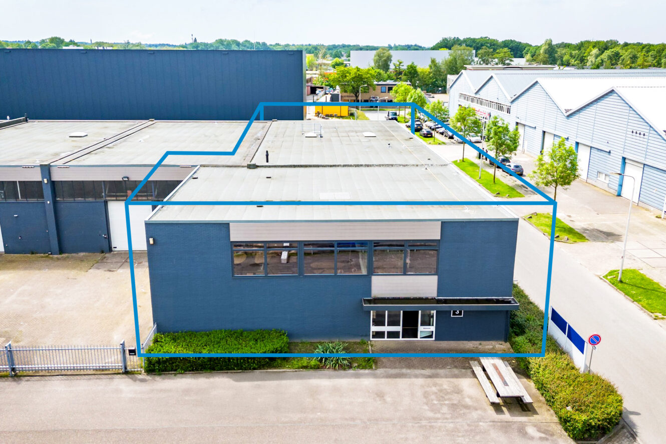 Luchtfoto van een commercieel industrieel gebouw met blauwe contouren, omringd door soortgelijke gebouwen en groen op de achtergrond.