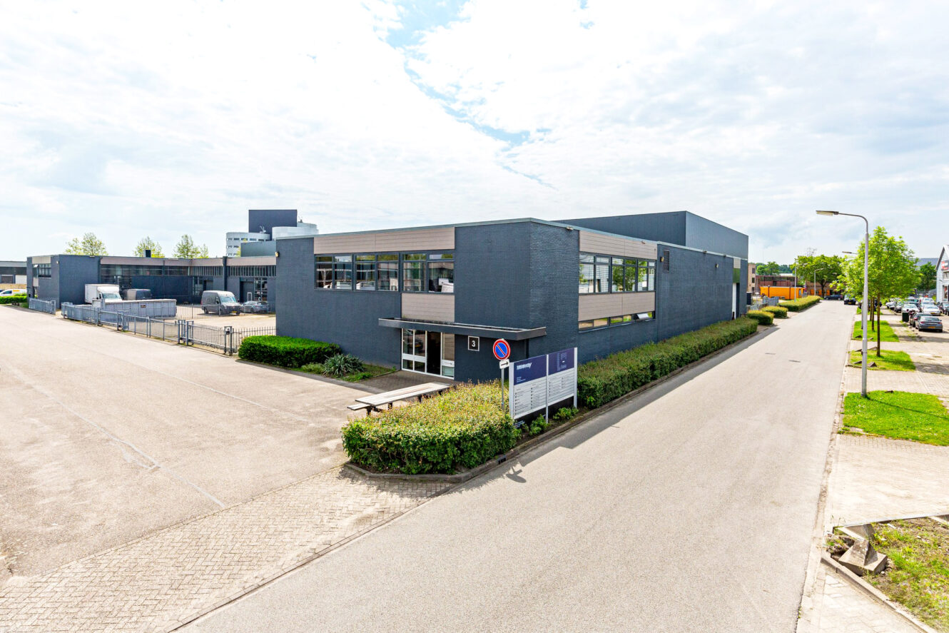 Modern industrieel gebouw met grijze buitenkant, grote ramen en een haag langs de omtrek, gelegen aan een rustige straat met een parkeerplaats en bewegwijzering bij de ingang.
