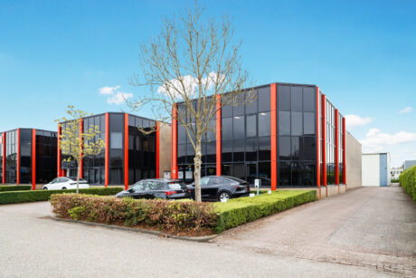 Modern kantoorgebouw met grote glazen ramen en rode accenten, omringd door geparkeerde auto's, gesnoeide heggen en een paar kleine bomen onder een strakblauwe hemel.