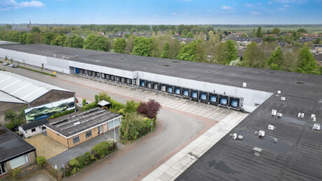 Luchtfoto van een groot industrieel magazijn met meerdere laadkades, een oprit en omliggend groen naast een woonwijk.