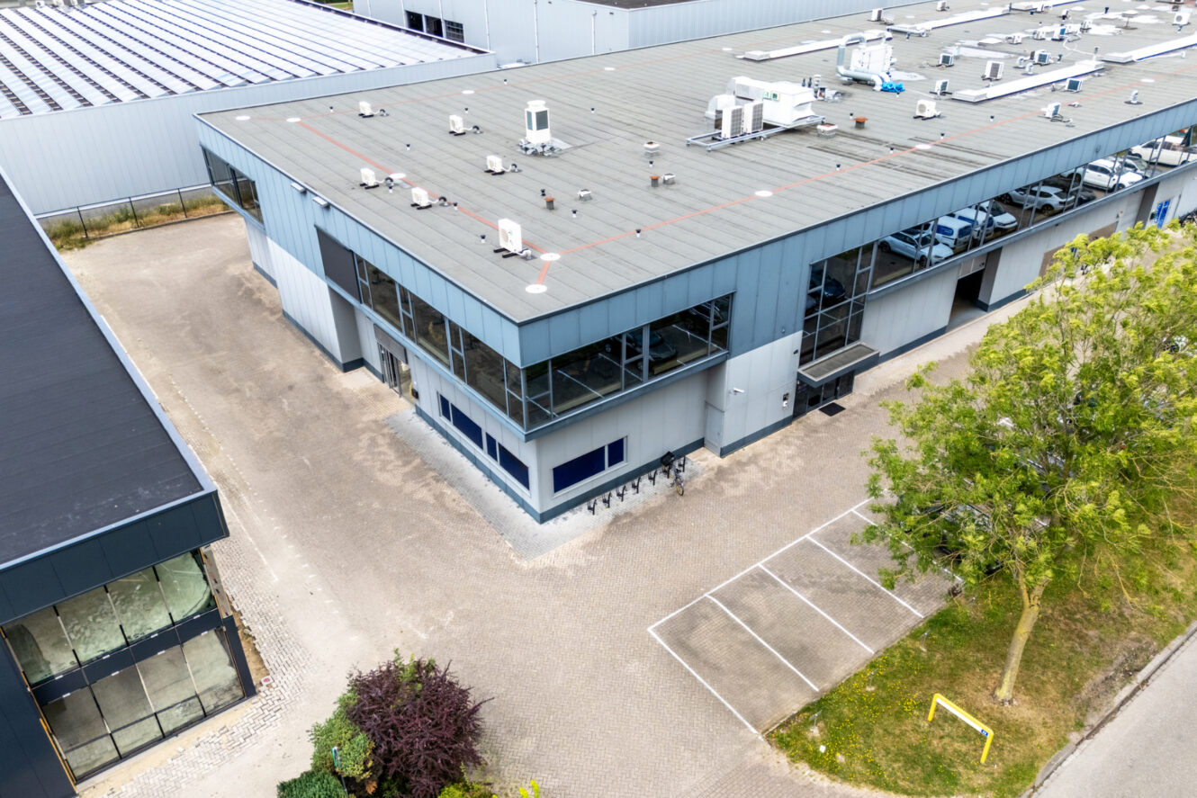 Luchtfoto van een modern industrieel gebouw met een plat dak, grote ramen, HVAC-units op het dak, parkeerplaatsen in de buurt en omringende bomen.