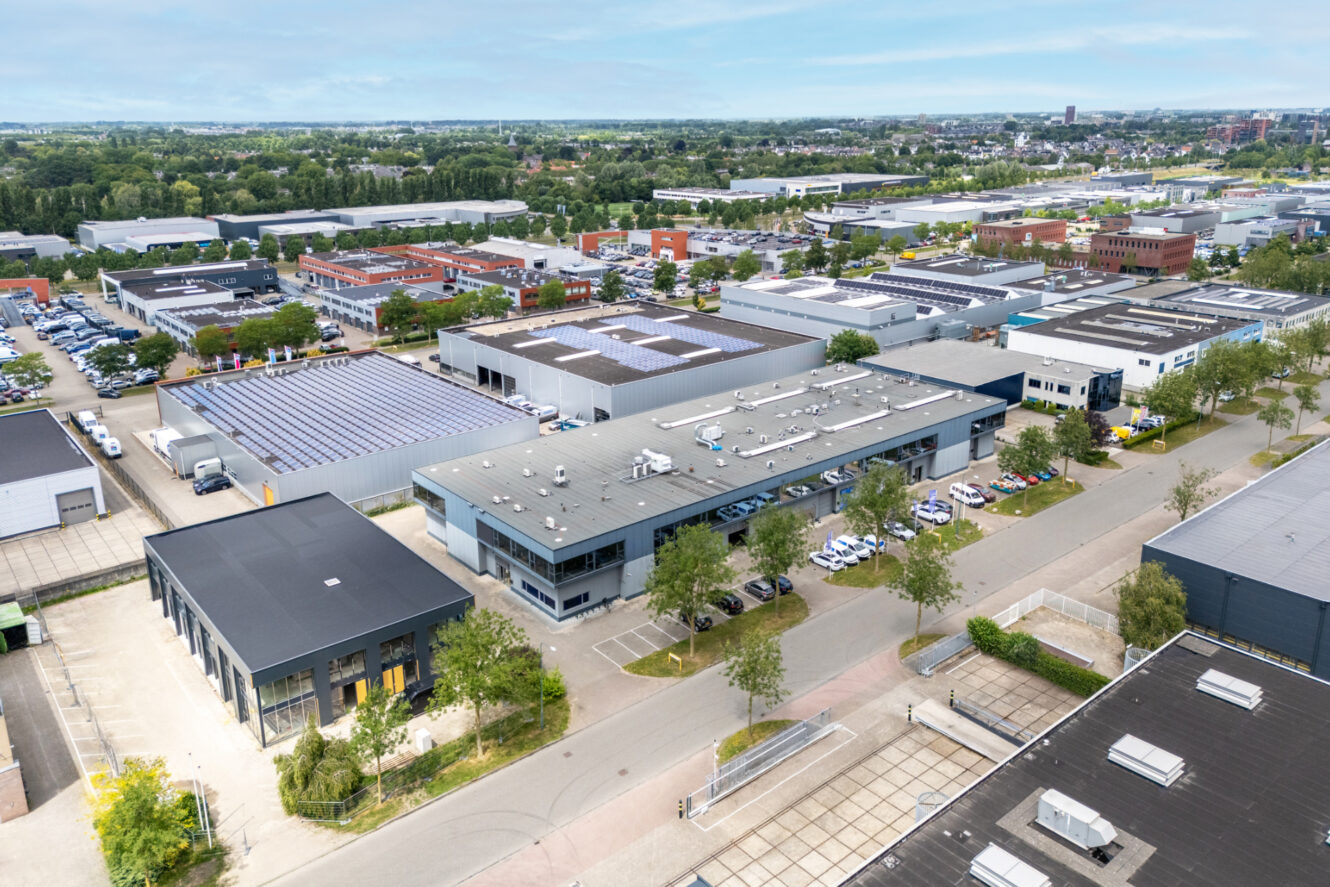 Luchtfoto van een industrieterrein met meerdere magazijnen, kantoorgebouwen, geparkeerde auto's en zonnepanelen op verschillende daken, omgeven door groen en wegen.