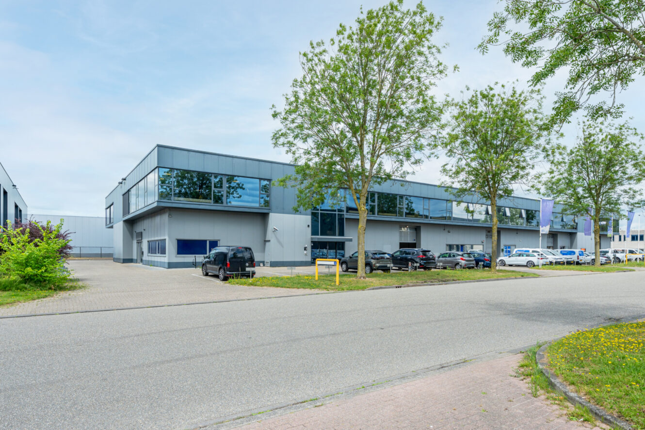 Modern industrieel gebouw met grote ramen, meerdere geparkeerde auto's en bomen langs de straat, gefotografeerd op een heldere dag.