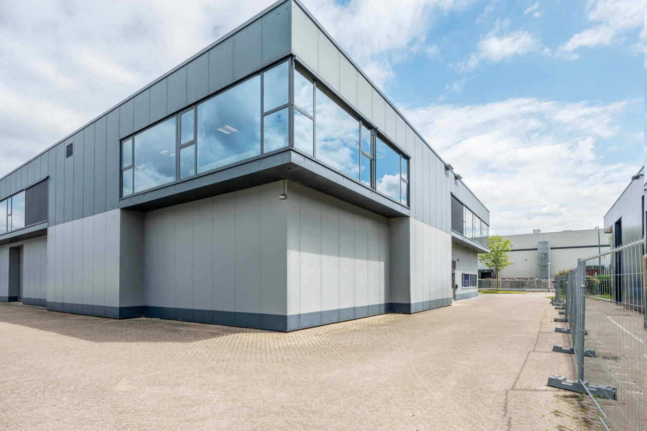 Een modern industrieel gebouw met grote reflecterende ramen, een grijze metalen buitenkant en een lege geplaveide binnenplaats onder een gedeeltelijk bewolkte hemel.