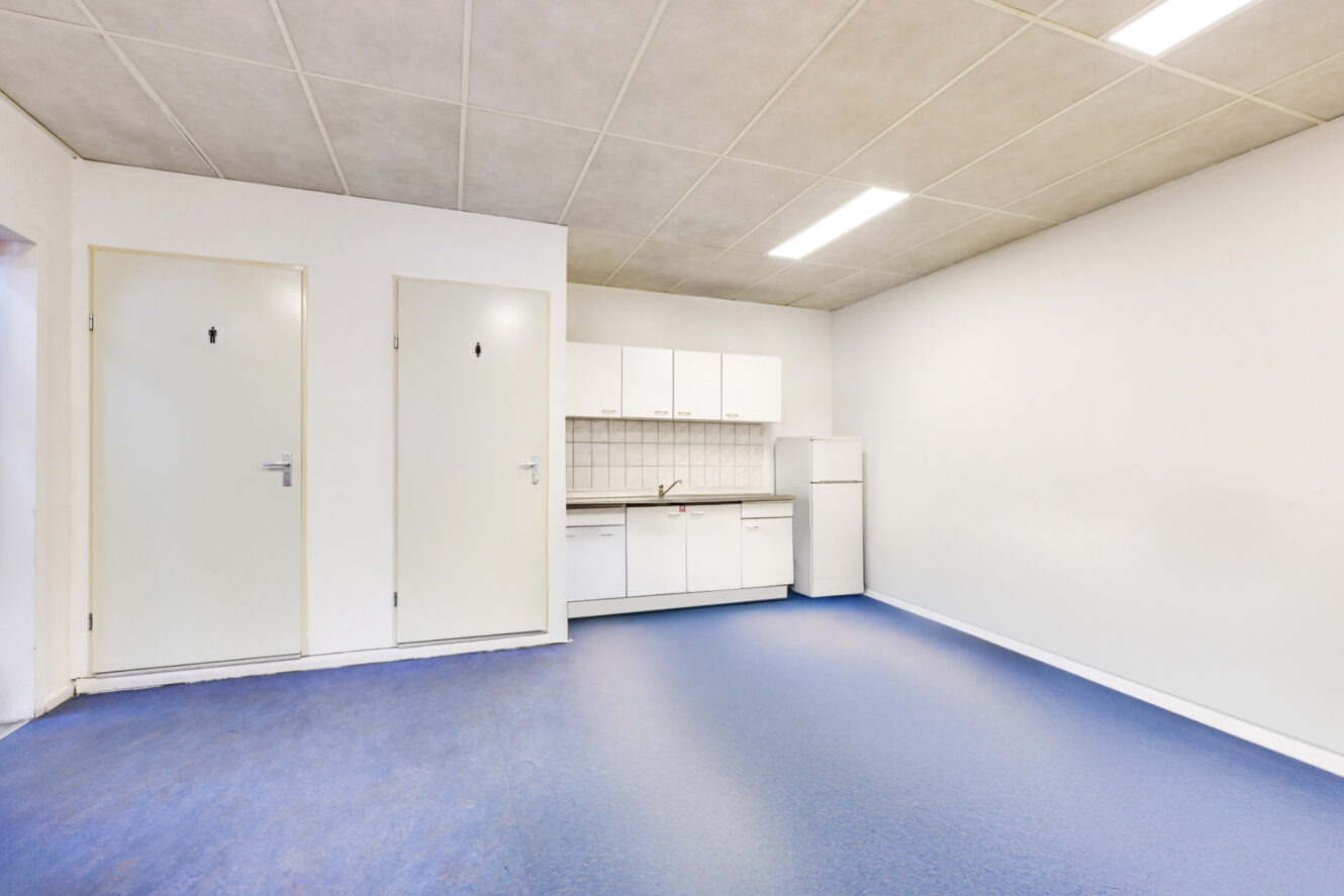 Een kleine kamer met blauwe vloer, witte muren, een keukentje met kastjes, een gootsteen, een koelkast en twee gesloten deuren met toiletsymbolen.