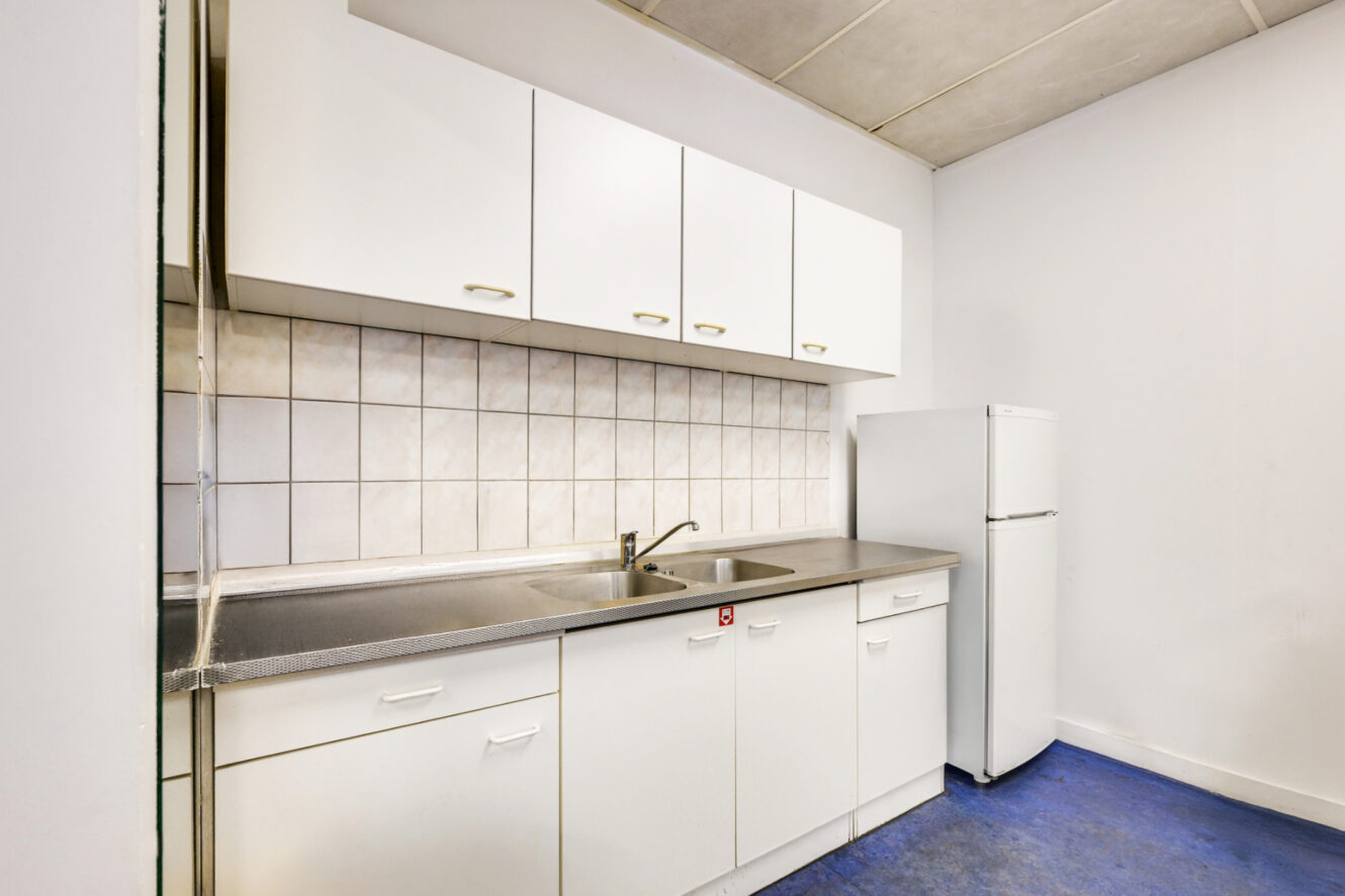 Een kleine keuken met witte kastjes, een roestvrijstalen gootsteen en aanrechtblad, een betegelde achterwand en een witte koelkast op een blauwe vloer.