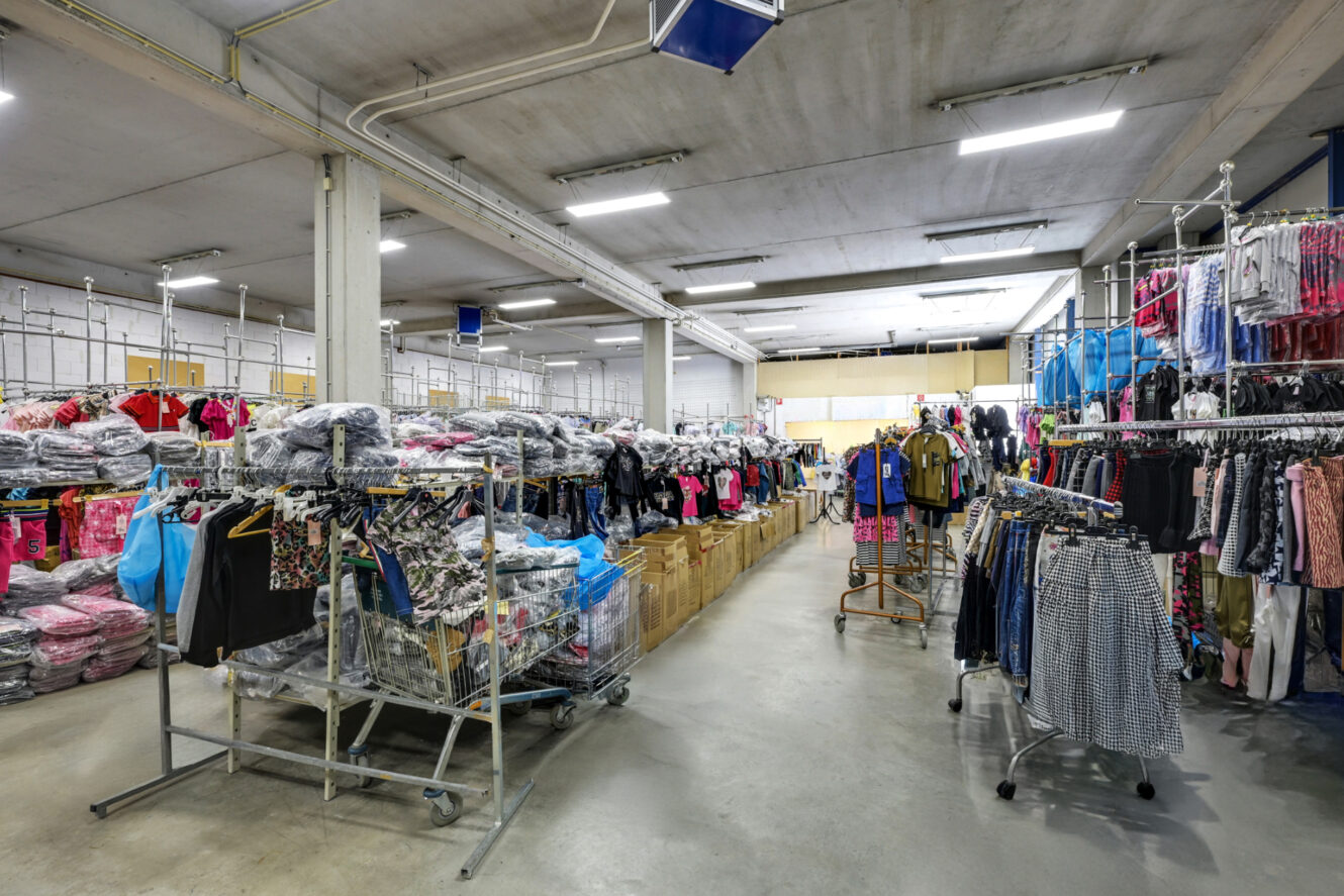 Groot magazijn met rekken en bakken vol met verschillende kledingstukken, heldere verlichting aan het plafond en betonnen vloeren.
