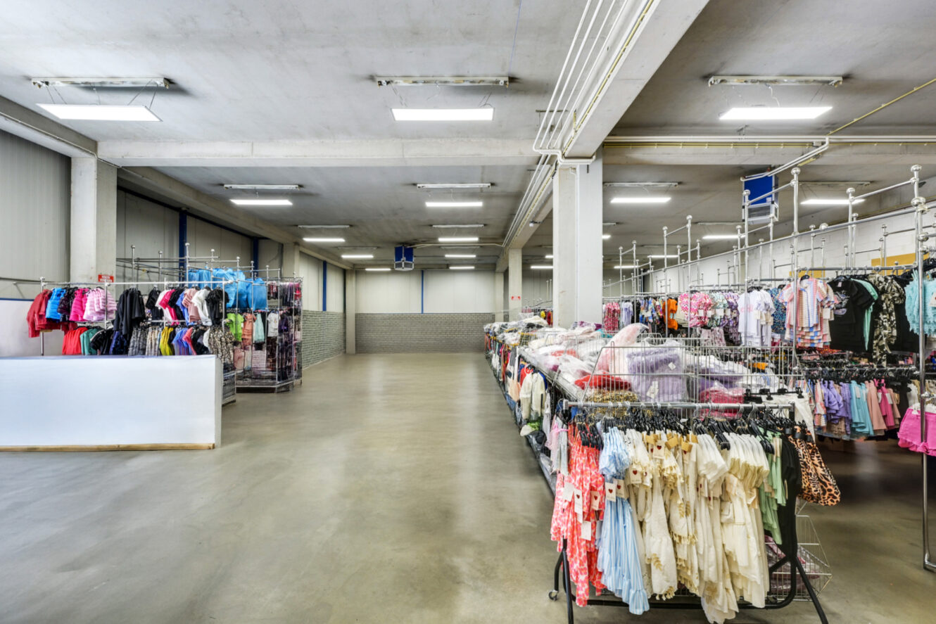 Een ruim kledingwinkelinterieur met rekken vol verschillende kledingstukken netjes gerangschikt onder fel fluorescerend licht.