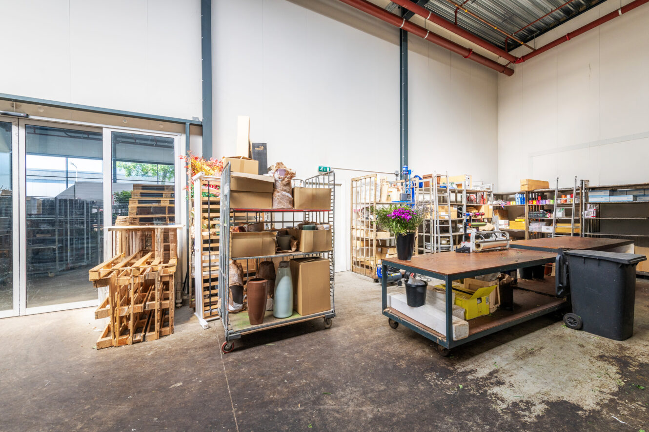 Een ruime industriële werkruimte met metalen planken, trolleys, houten pallets, vazen en benodigdheden georganiseerd tegen de muren; grote ramen en hoog plafond zichtbaar.