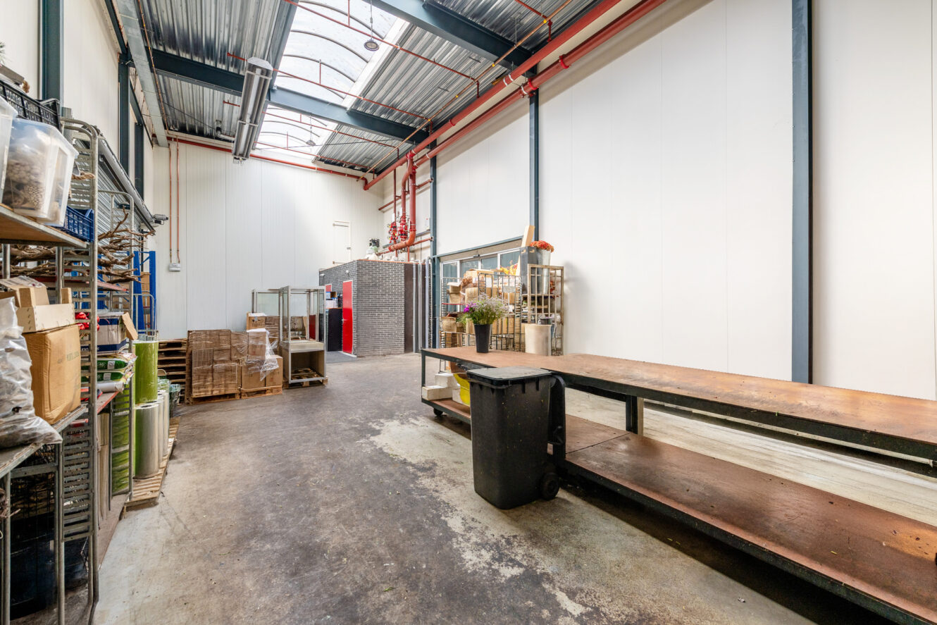 Industriële werkruimte met hoog plafond, metalen rekken, houten werkbanken, een prullenbak en verschillende benodigdheden netjes georganiseerd langs de muren. Natuurlijk licht valt binnen via dakramen boven.