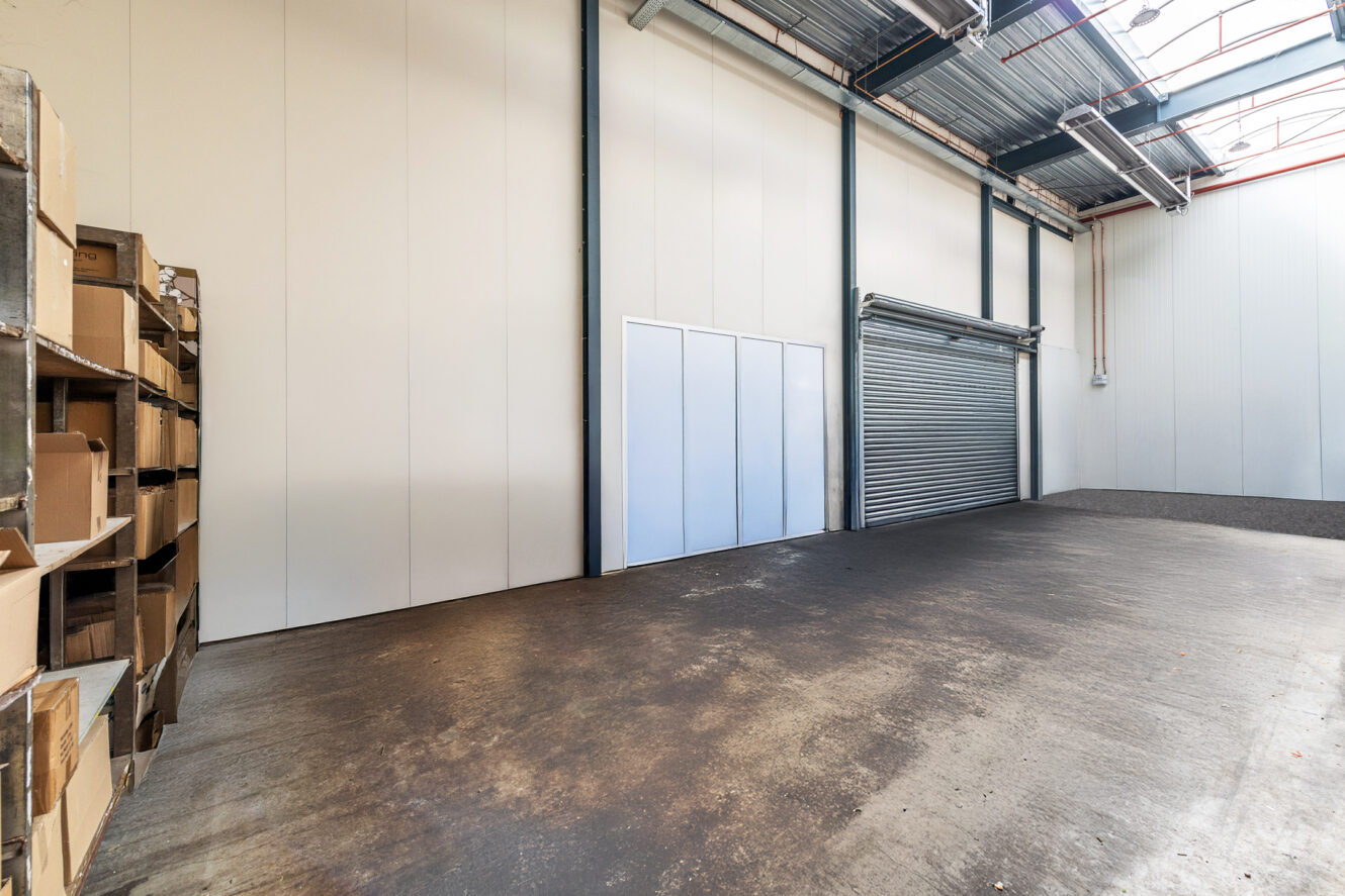 Leeg magazijninterieur met betonnen vloer, hoog plafond, metalen rekken aan de linkerkant en een grote gesloten roldeur aan de rechtermuur.