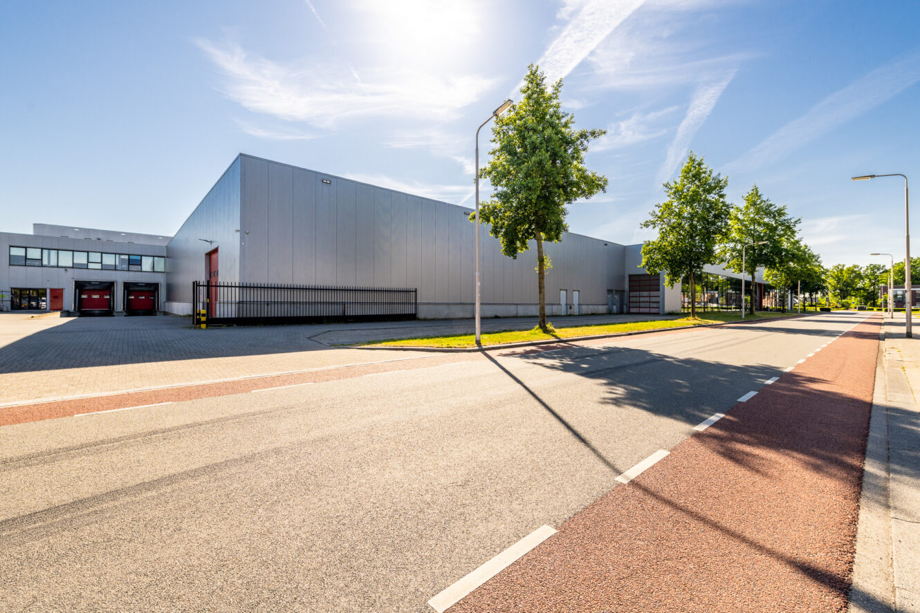 Groot modern magazijngebouw met rode laadperrons, gelegen aan een zonnige straat met bomen en een heldere hemel.