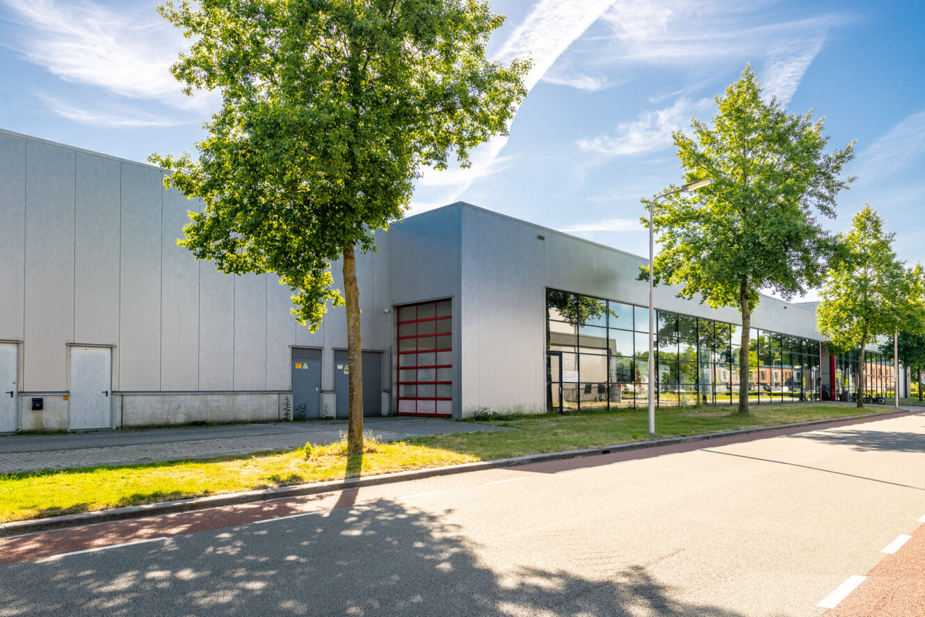 Modern industrieel gebouw met grote ramen en een rode garagedeur, gelegen langs een straat met bomen op een zonnige dag.