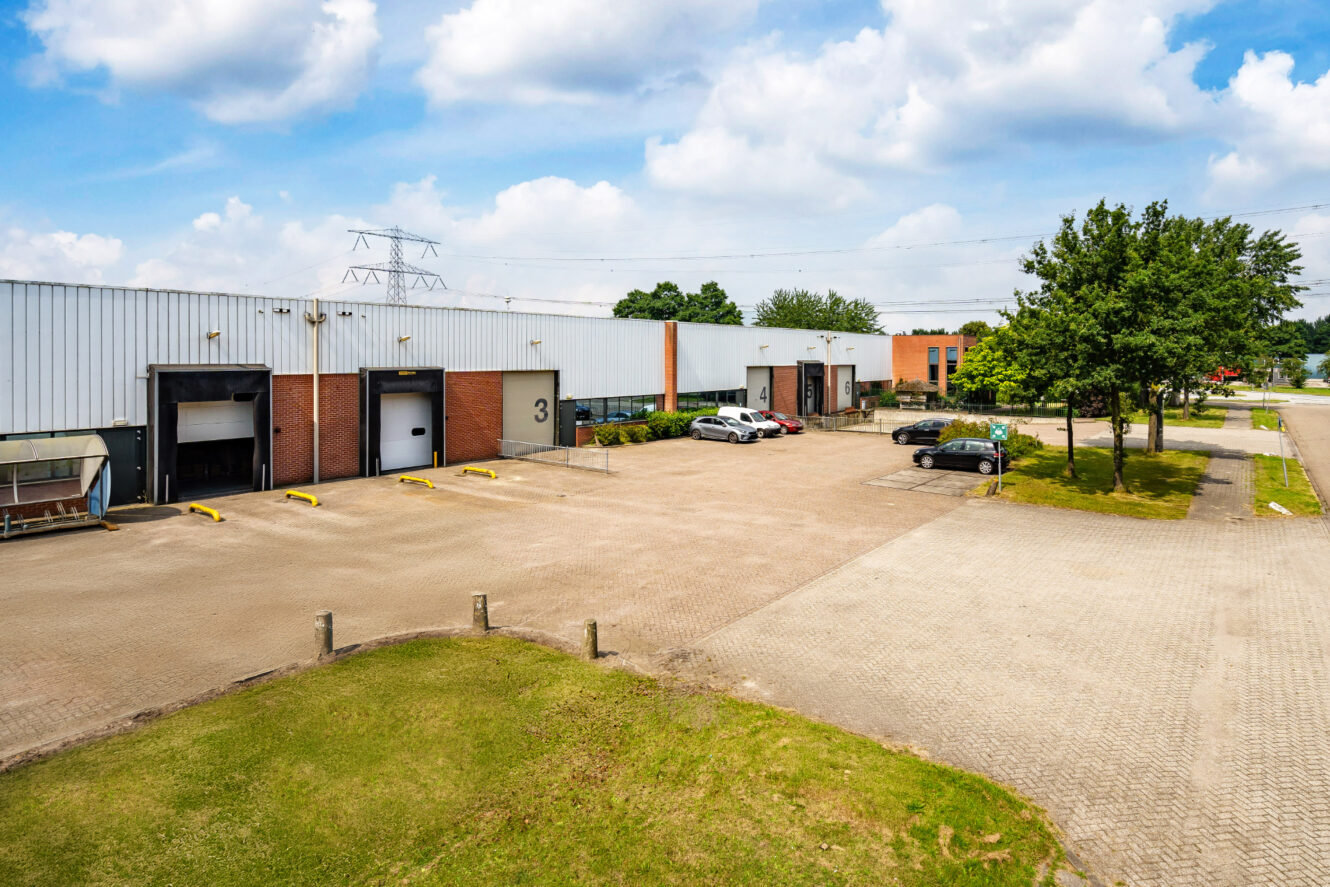 Industrieel pakhuis met meerdere laaddocks, een paar geparkeerde auto's en een verhard terrein omringd door bomen onder een gedeeltelijk bewolkte hemel.
