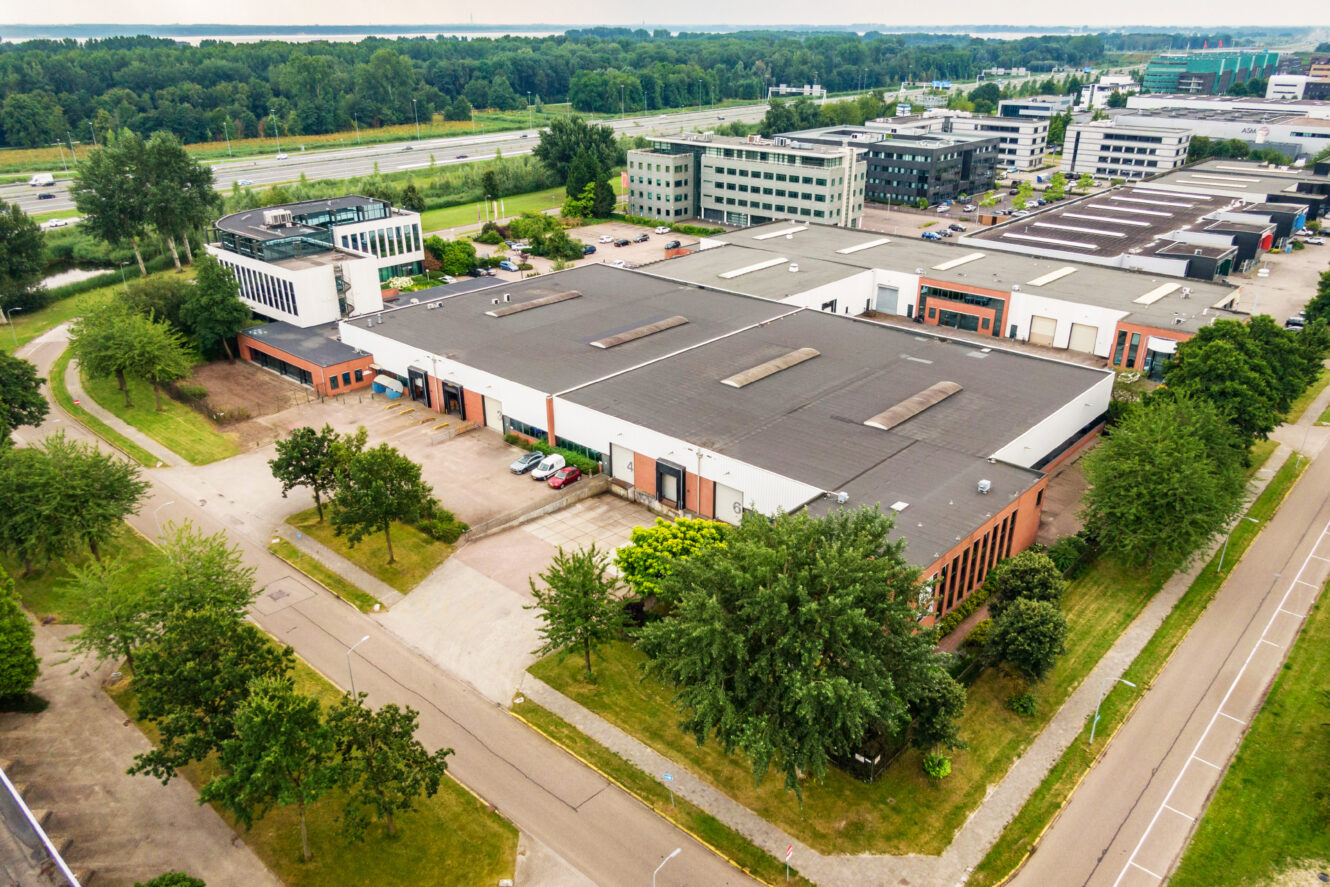 Luchtfoto van een industrieterrein met grote magazijngebouwen, parkeerplaatsen, omliggende wegen en groene bomen.