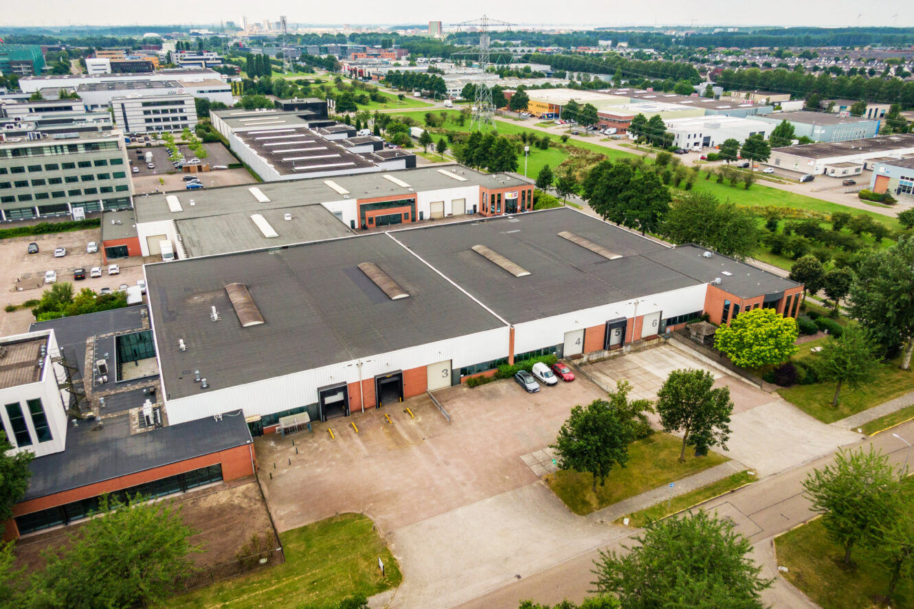 Luchtfoto van grote industriële magazijnen omringd door geparkeerde auto's, bomen en nabijgelegen kantoorgebouwen in een ontwikkeld gebied.