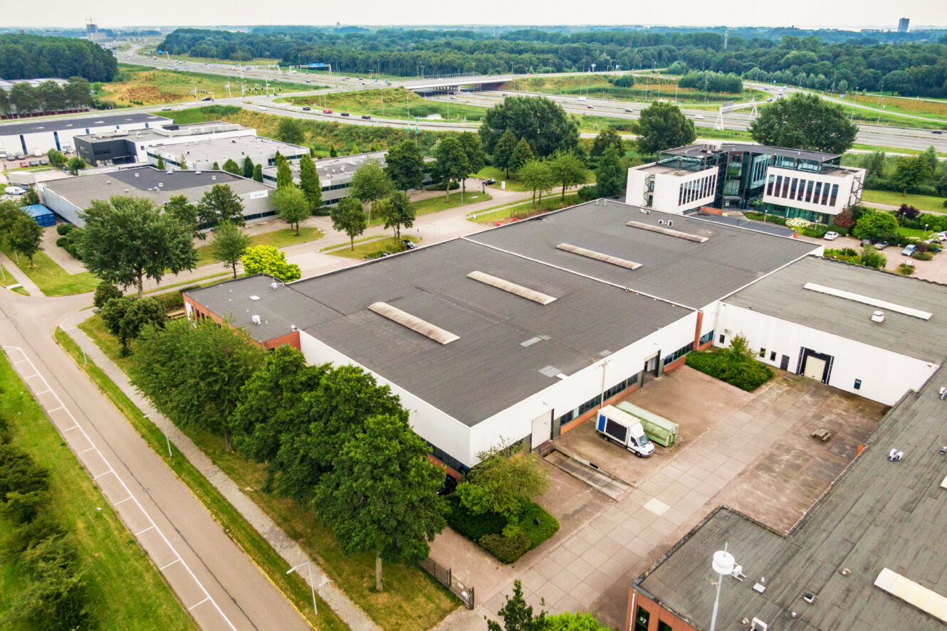 Luchtfoto van een groot industrieel pakhuis met een vrachtwagen buiten geparkeerd, omringd door bomen, wegen en nabijgelegen kantoorgebouwen.