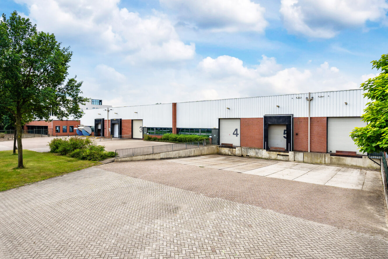 Een modern industrieel pakhuis met laaddocks, genummerde deuren en een verharde oprit, omringd door bomen en een bewolkte hemel.