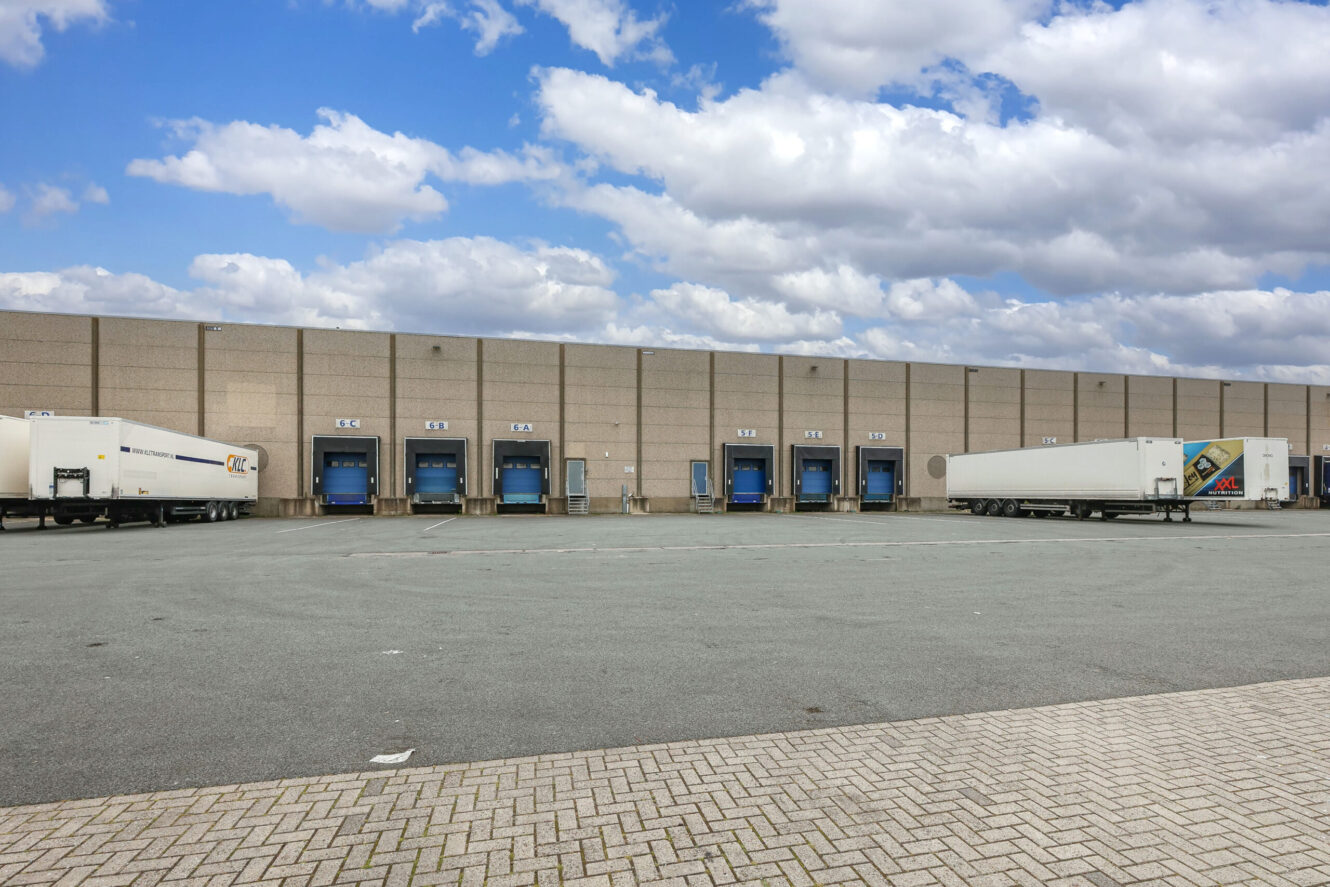 Breed uitzicht op een laadperron van een magazijn met verschillende blauwe laadperrons en geparkeerde opleggers onder een gedeeltelijk bewolkte hemel.